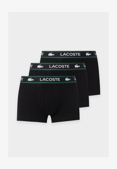 Trois paires de boxers noirs pour hommes avec des ceintures bordées de vert comportant le texte blanc Lacoste et les logos crocodile.