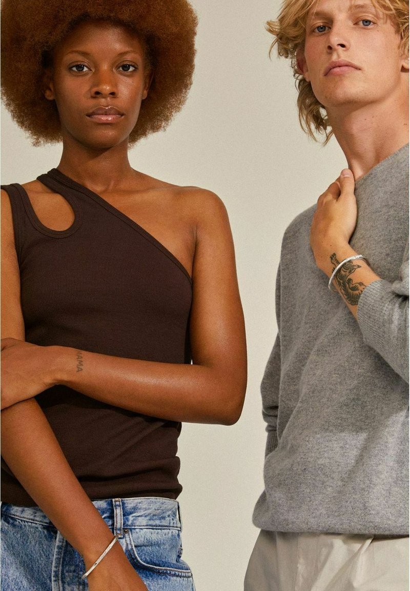 Top corto a coste marrone con design a spalla unica, abbinato a un maglione grigio a maglia. Entrambi i modelli indossano jeans casual e mostrano gioielli semplici.