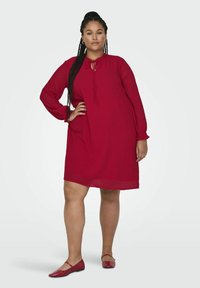 Czerwona sukienka do kolan z długimi rękawami, okrągłym dekoltem i detalem w postaci sznureczka. Gładka tekstura z subtelnym wzorem w kropki. Dopasowana z czerwonymi balerinkami.