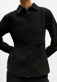 Camicia in denim nera con due tasche sul petto, chiusura con bottoni e colletto. Il tessuto ha una texture liscia con cuciture sottili.