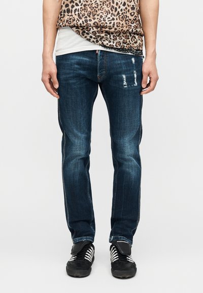 Homme portant un jean bleu foncé usé, des baskets noires à lacets blancs, et un haut imprimé léopard par-dessus une chemise blanche.