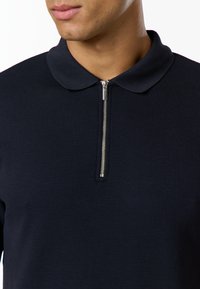 Homme portant une chemise texturée bleu marine foncé avec une courte fermeture éclair argentée et un col arrondi, visible du cou à la poitrine.