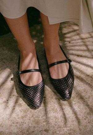 Kojos, dėvinčios juodus perforuotus Mary Jane stiliaus batus su sagtelės dirželiais ant betono grindinio, saulės šviesoje su šešėliais.