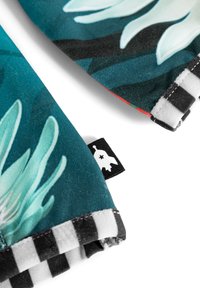 Tekstil med en teal baggrund og store, lyse blomster mønstre. Kanten har en sort-hvid ternet kant og et lille mærke.