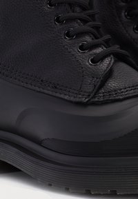 Gros plan d'une botte en cuir noir avec une surface texturée, des lacets noirs et une semelle en caoutchouc épaisse et rainurée.