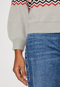 Ramię i ręka opierające się obok niebieskich jeansów, w szarej bluzie z szaro-czerwono-czarnym wzorem w zygzaki na mankiecie i w pasie.