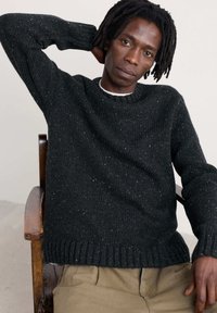 Crni pleteni pulover s teksturiranim uzorkom, okruglim izrezom i rebrastim manžetama i rubom. Nosite ga preko bež hlača, sjedeći na drvenoj stolici.