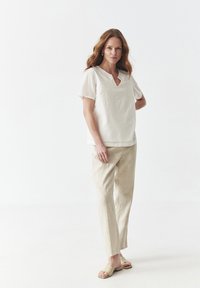 White casual blouse with short sleeves and embroidered front, paired with light beige linen pants and simple sandals, standing against a plain background.