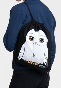 Black drawstring backpack featuring a white snowy owl graphic with brown eyes and yellow accents. Material appears sturdy and smooth.