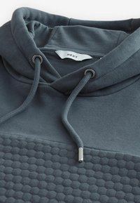Donkergrijze hoodie met een gestructureerd hexagonaal patroon aan de onderkant, verstelbaar trekkoord met metalen tips en zachte stof.