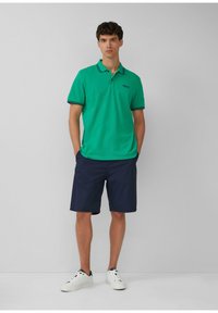 Grünes Poloshirt mit navyblauem gestreiften Kragen, kombiniert mit navyblauen Shorts. Das Outfit wird mit weißen Sneakers abgerundet.