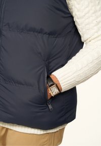 Person trägt einen weißen Strickpullover und beige Hose, hat die Hand in der Tasche einer marineblauen gesteppte Jacke und zeigt eine rechteckige goldene Armbanduhr.