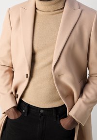 Abrigo de mezcla de lana color beige con solapas y botones, llevado sobre un cuello de tortuga color beige claro, combinado con jeans de mezclilla negros.