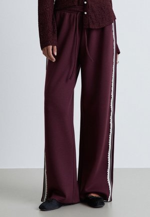 Personne portant un pantalon ample bordeaux avec des bandes blanches sur les côtés et une bordure en dentelle, associé à des chaussures plates noires et un haut bordeaux texturé.