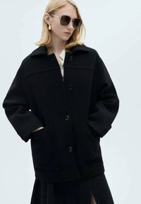Černá vlněná bunda s límečkem, velkými knoflíky, bočními kapsami a volným střihem. Model stojí před světlým pozadím.
