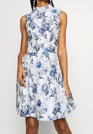 Biała sukienka bez rękawów z niebiesko-czarnym kwiatowym wzorem, dopasowana w talii i rozkloszowana spódnica, noszona przez modelkę z długimi kolczykami.