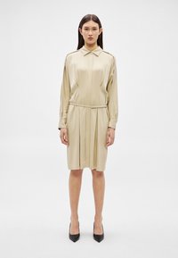 Žena stojaca čelom dopredu, oblečená v béžových dlhorukávových šatách s opaskom a v čiernych lodičkách s ostrou špičkou na jednoduchom bielom pozadí.