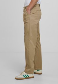 Chino din bumbac bej cu un design pe picior drept, având buzunare laterale și un buzunar la spate, asortați cu adidași albi și verzi.