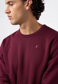 Felpa a maniche lunghe color bordeaux in misto cotone, con scollo a girocollo, colletto a costine e un piccolo logo ricamato sul petto.