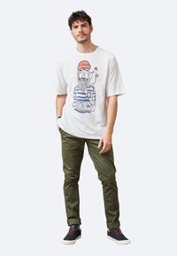 Camiseta blanca de manga corta con un diseño de rayas azules que presenta una impresión gráfica de un hombre con barba y un sombrero rojo. Pantalones verde oliva y zapatos oscuros.