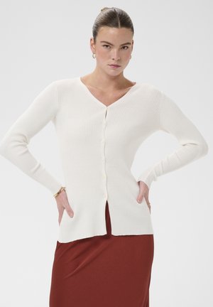 Femme avec les cheveux attachés en chignon portant des boucles d'oreilles créoles dorées, un cardigan blanc côtelé boutonné et une jupe marron, posant les mains sur les hanches.