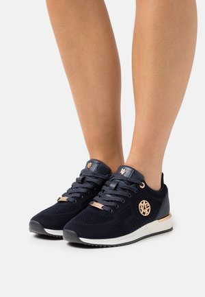 Piedi che indossano scarpe da ginnastica in mesh blu navy scuro con accenti in oro rosa e suole intermedie bianche su uno sfondo chiaro uniforme.