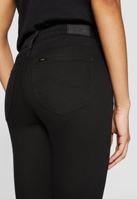 Jeans en denim noir avec une coupe ajustée. Les caractéristiques incluent une ceinture en cuir, des poches arrière courbées et des détails de couture discrets.