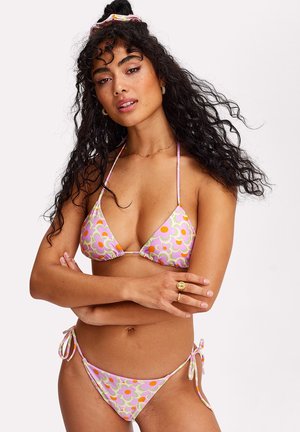 Femme aux longs cheveux bouclés portant un bikini à fleurs rose et un chouchou assorti, debout les bras croisés devant un fond blanc.