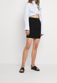 Falda de mezclilla negra, estilo ajustado, hasta la rodilla. Combinada con una camisa anudada de color azul claro y sandalias negras con tiras y suela plana.