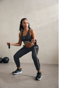 Crni sportski crop top i sužene hlače, držeći trake za vježbanje. Obuća ima crne, zelene i bijele naglaske. Neutralna pozadina.