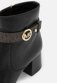 Bottine en cuir noir avec talon bloc, présentant une sangle marron à motif et une boucle dorée avec le logo MK.