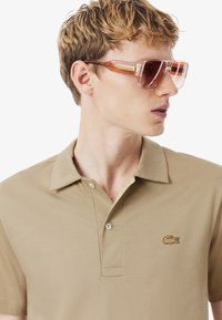 Beige pikétröja med krage och en subtilt broderad logotyp. T-shirten har en slät textur och har två knappar vid halsringningen.