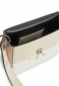 Sac Calvin Klein en cuir synthétique beige et blanc. L'intérieur comprend une doublure noire, une fermeture carrée et un emplacement pour carte. Bandoulière réglable incluse.
