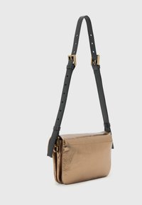 Metallisk bronsefarget crossbody-veske med svart justerbar stropp, med gullfargede metallbeslag og glatt tekstur. Kompakt og rektangulær form.