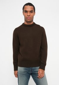 Brun stickad tröja med ribbat rundhals, långa ärmar och avslappnad passform, bärs med urtvättade blå jeans.