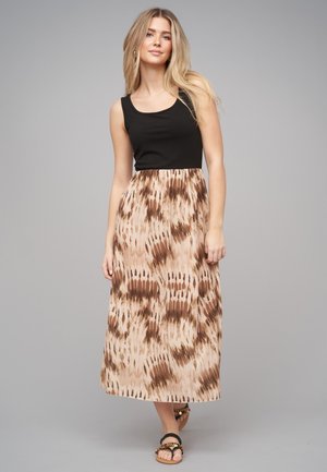Vrouw met lang blond haar, gekleed in een mouwloze zwarte top en een beige midi-rok met patroon, staand tegen een effen grijze achtergrond.