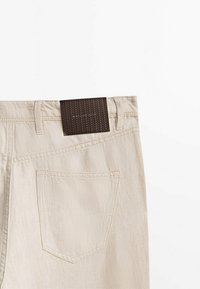 Beige puuvillapantalonit, joissa on kudottu ruskea nahkainen bränditunnus vyötäröllä. Sisältää yhden takataskun ja hienovaraisia ompeluyksityiskohtia.