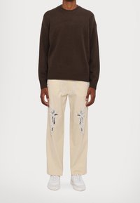 Brown knit sweater with ribbed cuffs paired with beige trousers featuring silver palm tree embroidery and white sneakers.