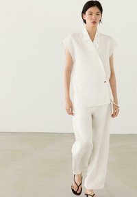 Femme portant un chemisier blanc sans manches avec un bouton, un pantalon large blanc et des sandales noires, debout sur un sol et un mur clairs unis.