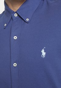Blå skjorta med knappar och vita knappar samt en vit broderad polo-spelare logo på vänster bröst.