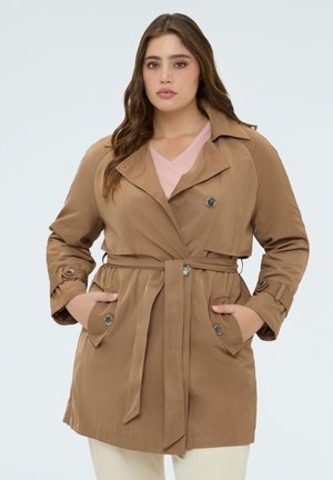 Mujer con un abrigo marrón abotonado con cinturón atado, manos en los bolsillos y una blusa de color rosa claro debajo.