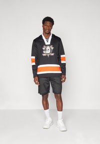 Juodas ledo ritulio marškinėliai su oranžinėmis ir baltomis juostelėmis, su centriniu logotipu; derinami su juodais tinkliniais šortais ir baltais sportbačiais.