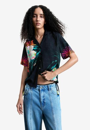 Woman with curly hair wears black short-sleeve shirt with colorful floral print and loose light blue jeans against white background.