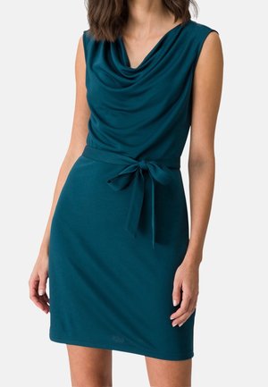 Sleeveless teal dress with cowl neck and matching fabric belt tied at waist, worn by a woman with loose, shoulder-length hair.