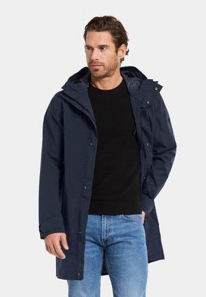 Mann trägt einen marineblauen Regenmantel mit Kapuze über einem schwarzen Pullover und blauen Jeans, steht mit einer Hand in der Tasche und blickt zur Seite.
