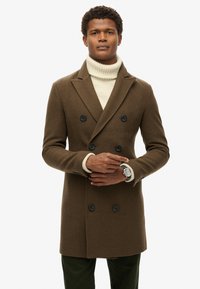 Dobbeltbrystet brun uldfrakke med store sorte knapper, båret over en cremet turtleneck. Modellen viser et ur på håndleddet.