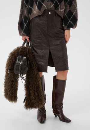 Elle porte une jupe en cuir marron foncé à motif crocodile et des bottes, tenant un sac assorti avec un manteau en fourrure marron à poils longs drapé sur le bras.