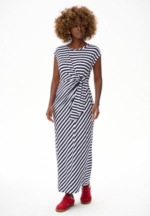 Kobieta o kręconych włosach, ubrana w granatowo-białą maksi sukienkę w paski z węzłem z boku i czerwone trampki, stojąca na białym tle.