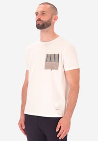 T-shirt bianca a maniche corte con una tasca a toppa testurizzata caratterizzata da strisce verticali blu, marroni e beige. Materiale in cotone morbido.