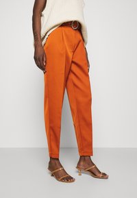 Oranje op maat gemaakte broek met een hoge taille, scherpe vouwen en zijzakken, gecombineerd met tan kleurige sandaaltjes met bandjes. Gebreid off-white sweater bovenaan.
